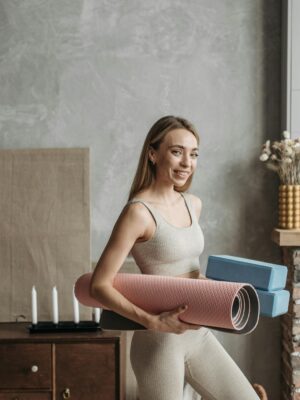 A smiling woman in activewear holding a yoga mat and blocks in a stylish home interior.
