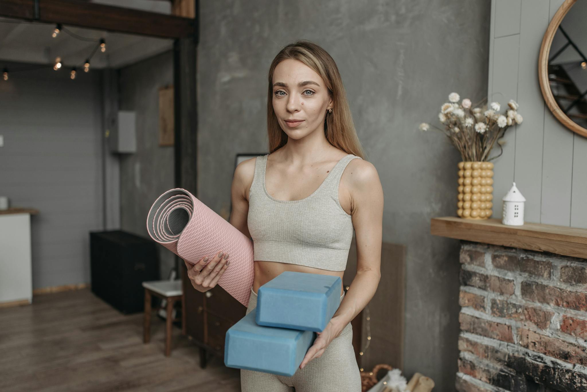 A woman in a sports bra holds yoga mat and blocks, standing in a modern studio setting.